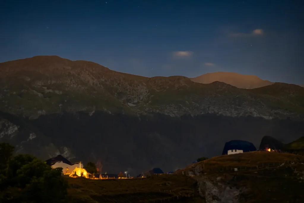 Noćni pogled na selo Lukomir pod planinom Bjelašnicom, s kućama osvijetljenim toplim svjetlom i planinskim vrhovima obasjanim mjesečinom.